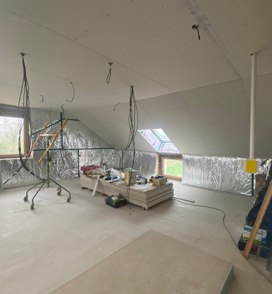 Pièce en rénovation avec murs recouverts de plastique, outils suspendus et établis sous un plafond mansardé