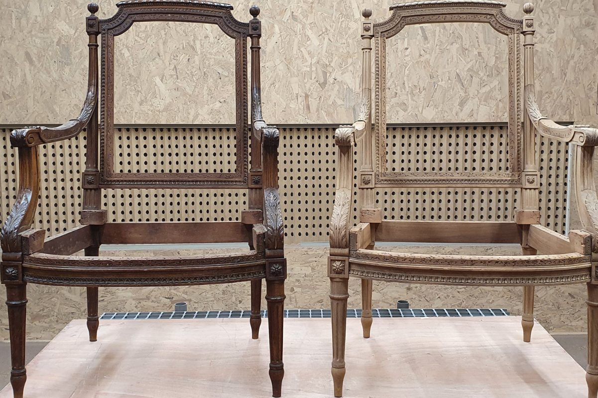 Deux chaises en bois finement ouvragées, l'une foncée et l'autre claire, sont disposées côte à côte devant un mur texturé.