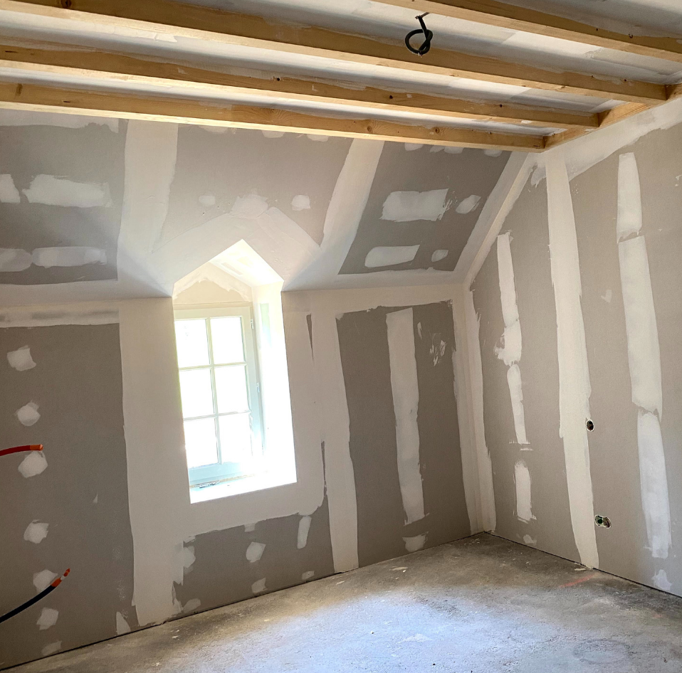 Pièce en plaques de plâtre inachevées avec plafond en pente, poutres apparentes, petite fenêtre et sol en béton brut.