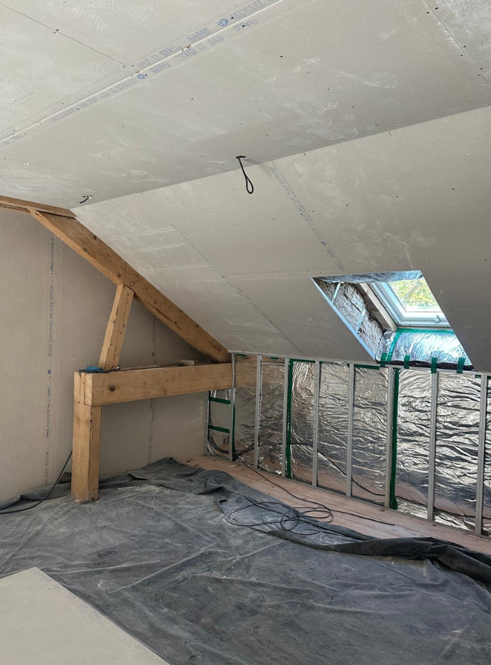 Pièce mansardée en cours d'aménagement avec poutres apparentes, isolation, bâches en plastique et fenêtre de toit inclinée