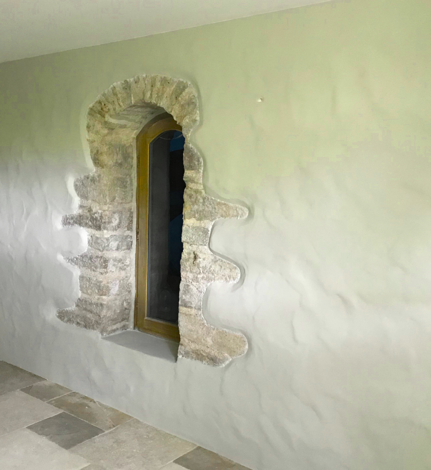 Mur intérieur avec une petite fenêtre cintrée encadrée de pierre apparente et de plâtre blanc.