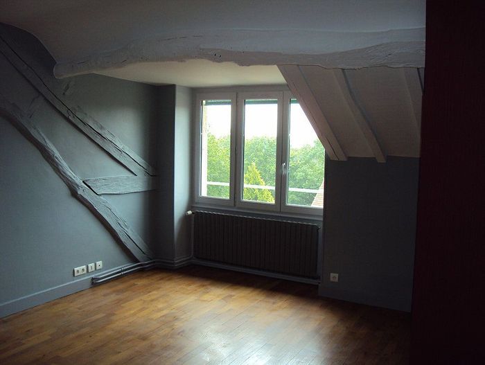 Grenier vide aux murs sombres, plafond mansardé, parquet et fenêtre donnant sur des arbres.