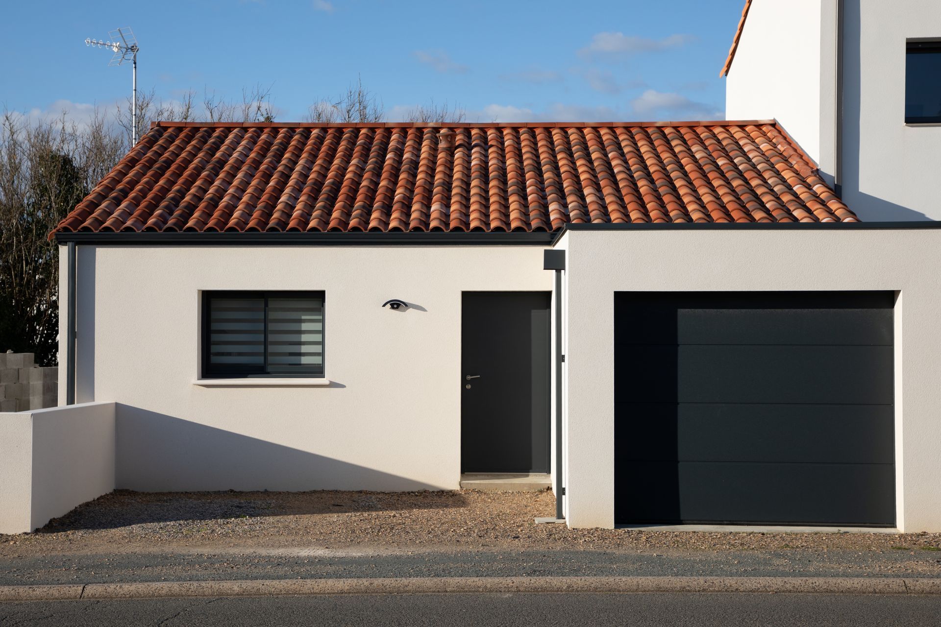 Petite maison blanche au toit de tuiles rouges et à la porte de garage sombre, donnant sur une allée de gravier.