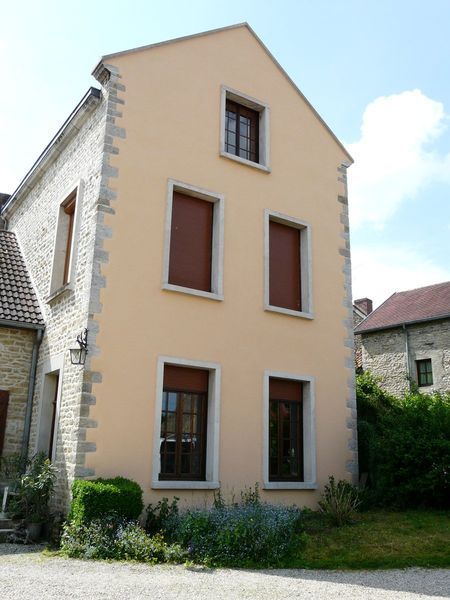 Maison beige de trois étages avec des ornements en pierre et des volets bruns, donnant sur une cour gazonnée.