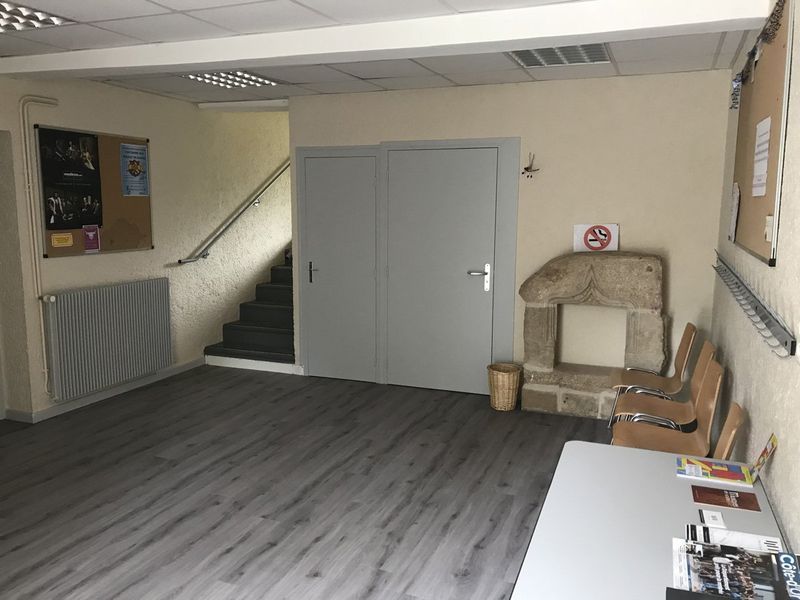 Hall d'entrée vide avec sol gris, escalier, porte fermée, radiateur et bureau blanc.