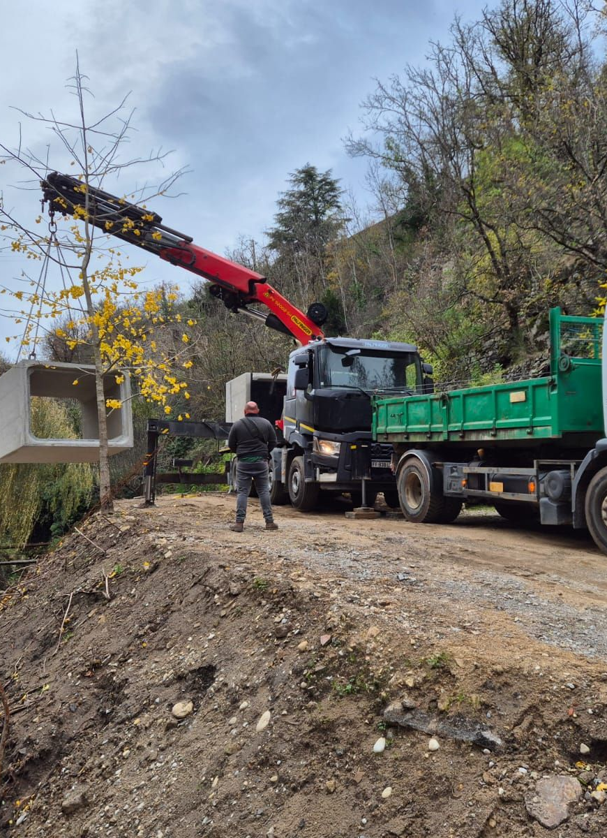 Une grue soulève une structure en béton ; deux camions circulent sur un chemin de terre à flanc de colline.