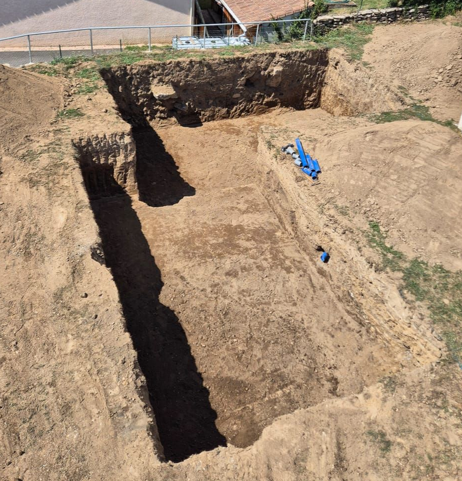 Tranchée rectangulaire creusée dans de la terre brune, probablement un site de fouilles archéologiques, à l'extérieur, près de bâtiments.
