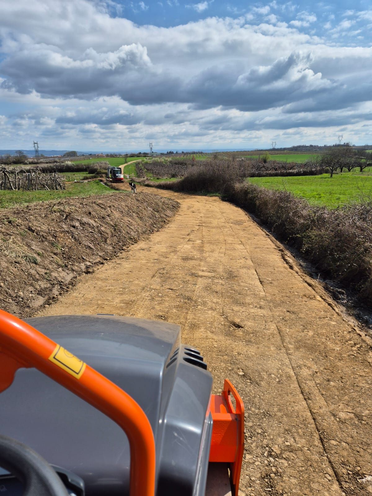 Une machine compacte un chemin de gravier serpentant à travers un paysage rural sous un ciel nuageux.