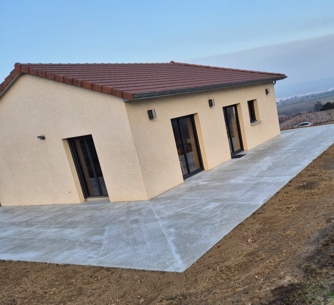 Maison beige au toit de tuiles rouges, posée sur une grande dalle de béton. Le paysage se dessine au loin.
