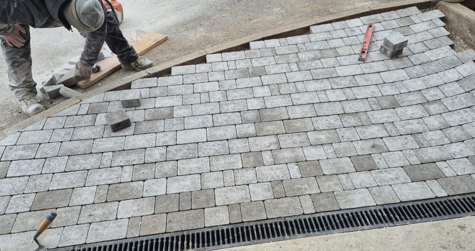 Un ouvrier du bâtiment pose des pavés sur une surface courbe, à proximité d'un drain.