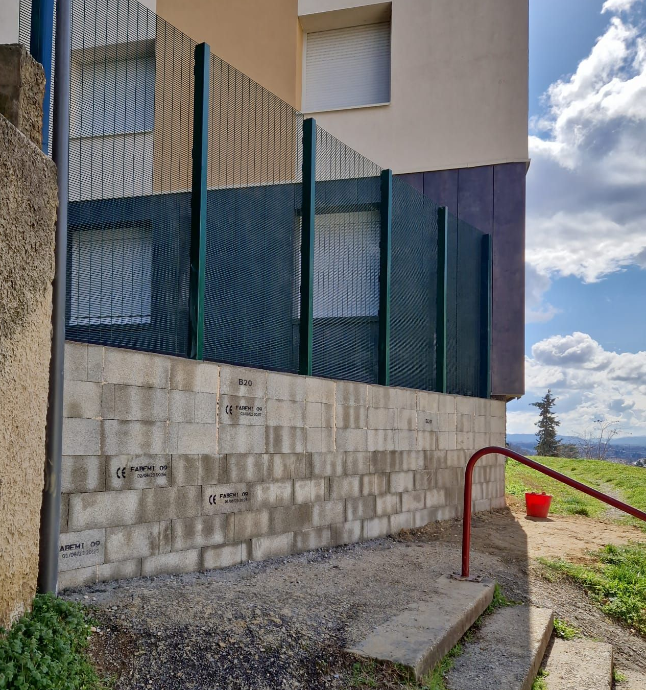 Clôture verte au-dessus d'un mur en parpaings, à côté d'un bâtiment. Une rampe et des marches rouges se trouvent à droite.