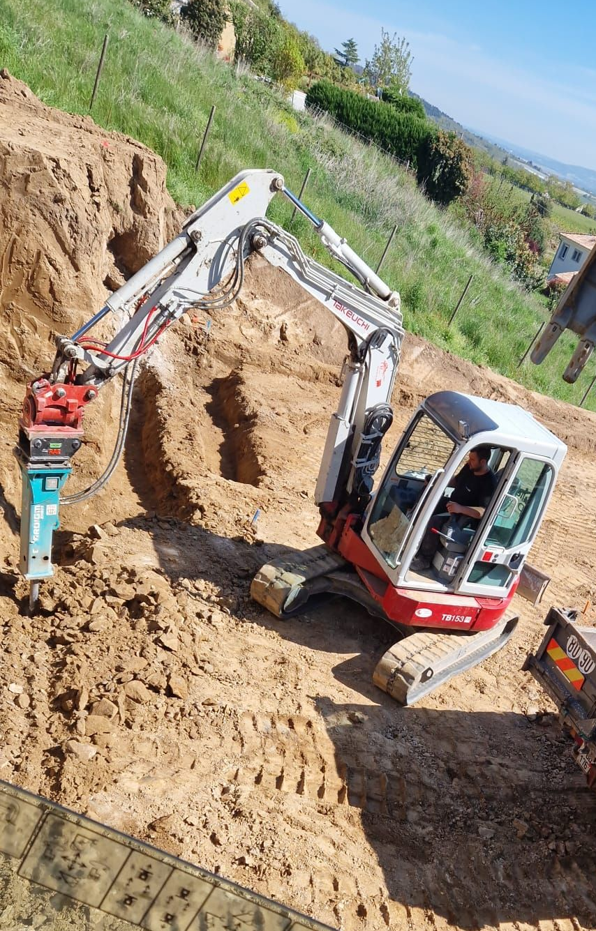 Pelle compacte équipée d'un marteau-piqueur en action sur un flanc de colline, autres engins à proximité.