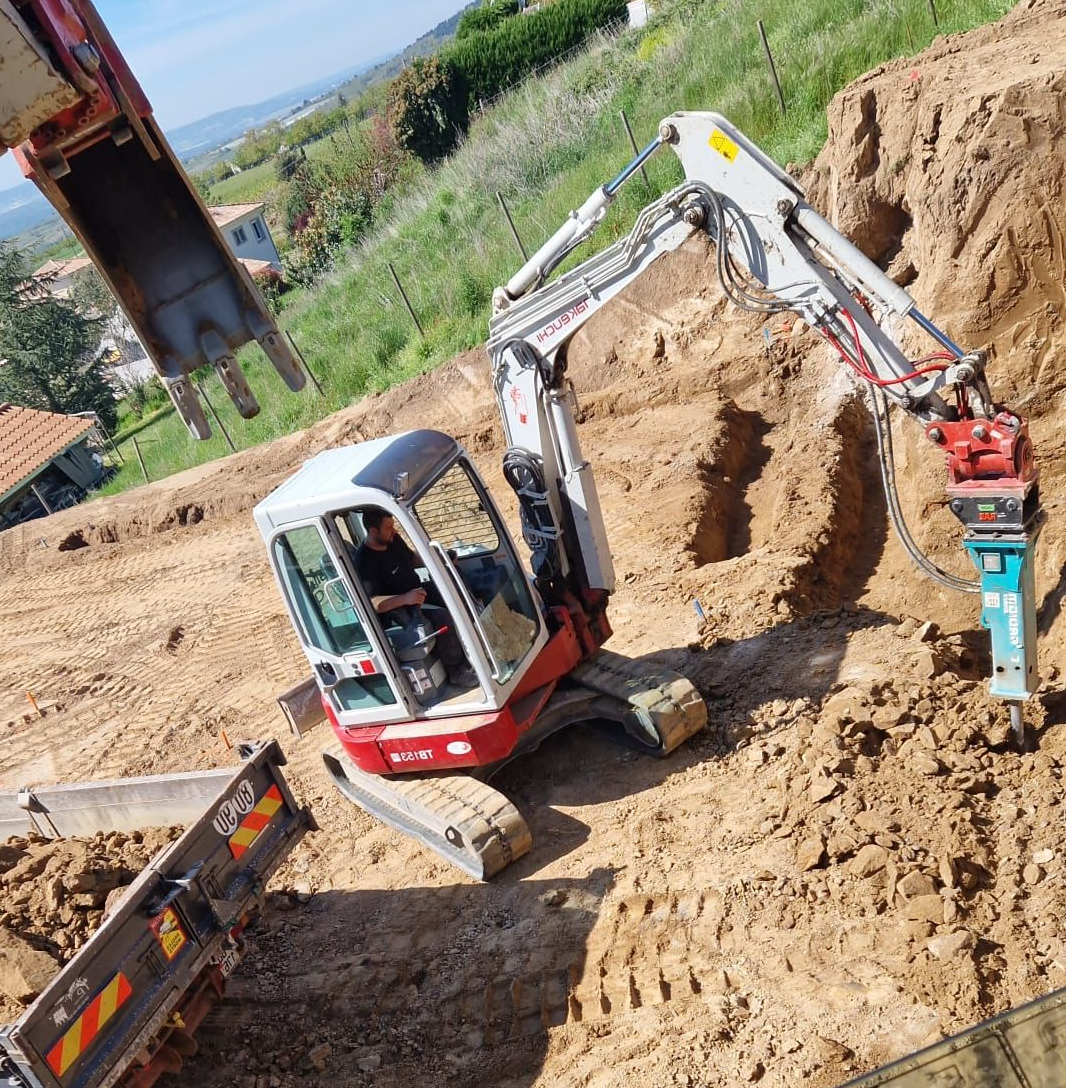 Une excavatrice équipée d'un marteau-piqueur travaille dans une fosse à terre, à côté d'un petit camion, dans un environnement extérieur vallonné.