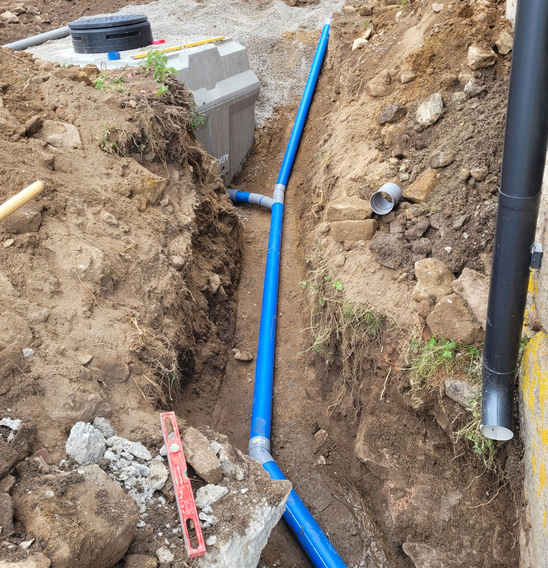 Un trou rectangulaire creusé dans une cour herbeuse, préparé pour accueillir une construction, entouré de terre et de piquets.