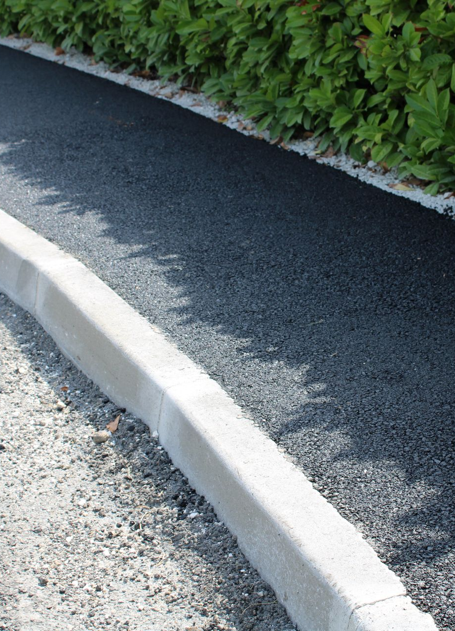 Chemin asphalté bordé d'un trottoir en béton et de gravier, avec une haie sur le côté.