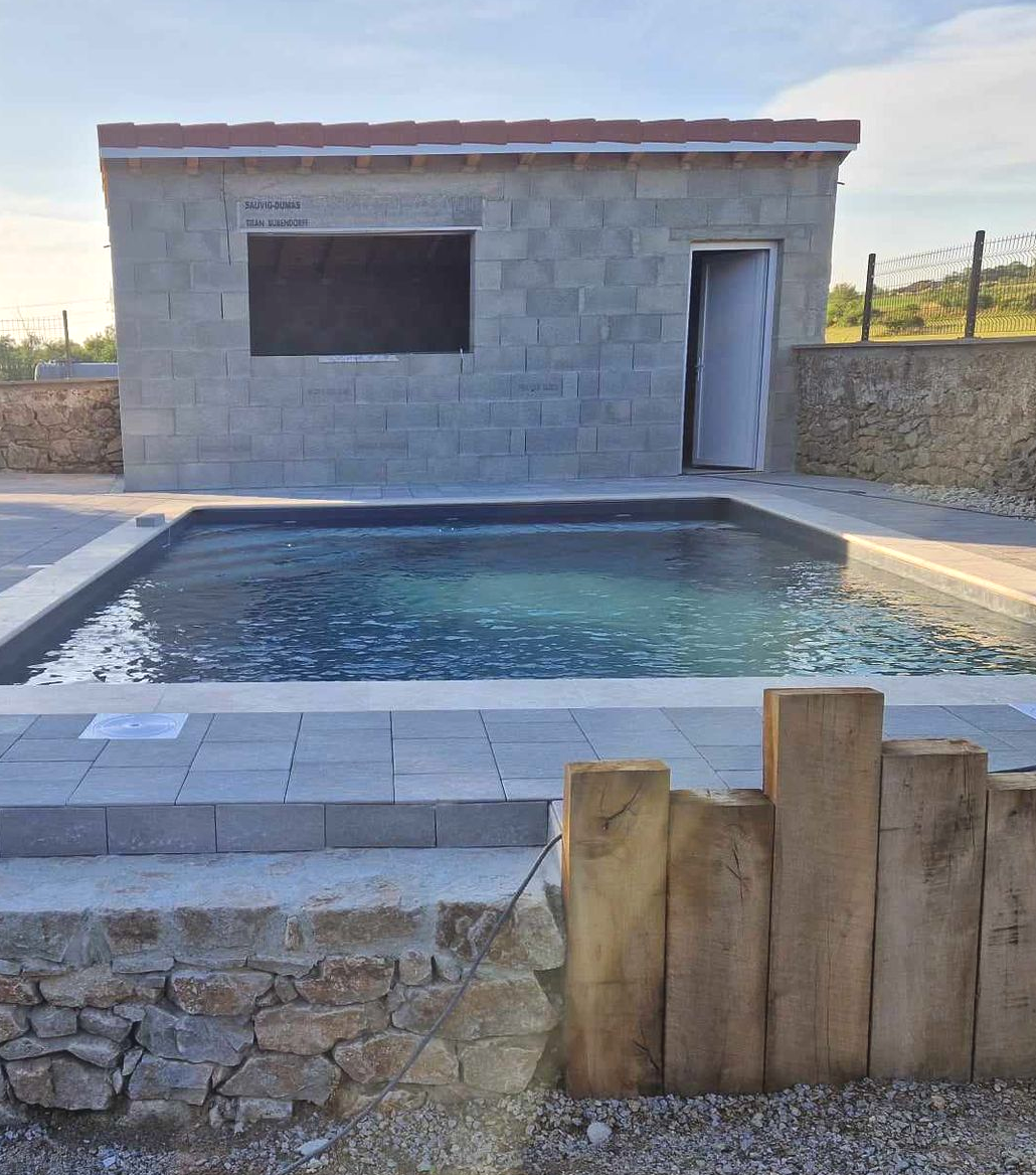 Piscine avec un bâtiment en béton inachevé, des murs en pierre et des poteaux de clôture en bois sous un ciel bleu.