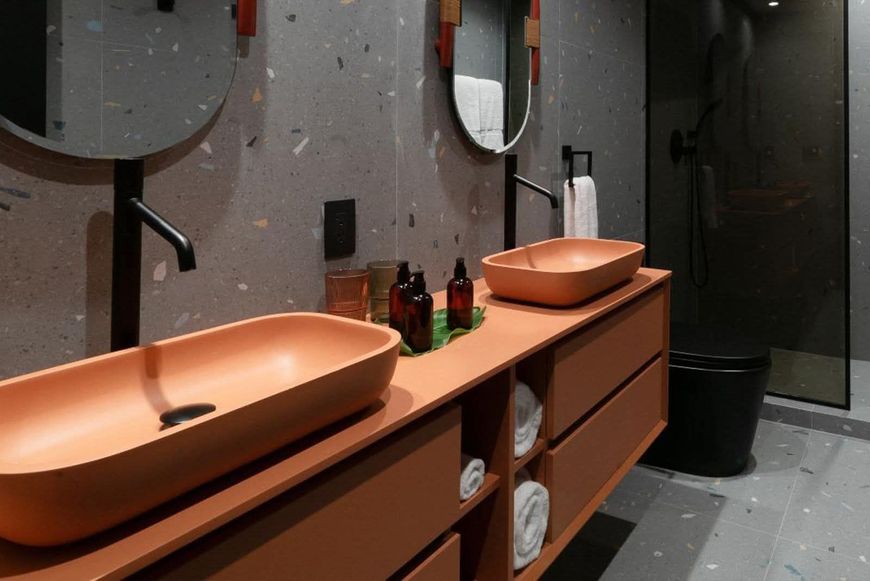 Baño con lavabos de color coral, armarios, grifería oscura, espejos circulares y paredes grises moteadas.