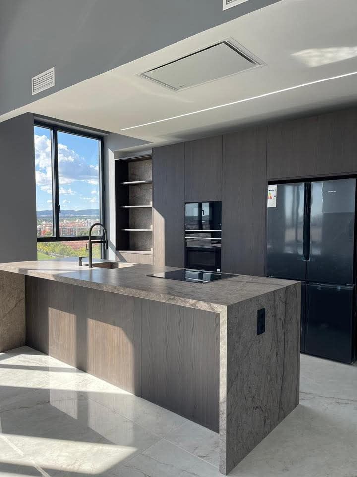 Cocina moderna con gabinetes grises, isla y ventana grande; electrodomésticos empotrados.