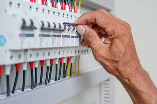 Hand flipping a circuit breaker switch in an electrical panel, colored wires visible, indoor setting.