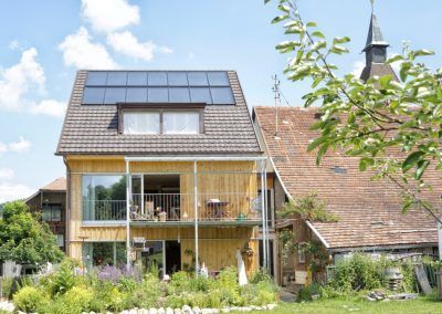 Modernes Haus mit Sonnenkollektoren neben einem alten Gebäude. Grüne Pflanzen im Vordergrund.