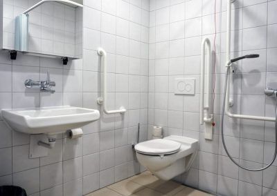 Barrierefreies Badezimmer mit weißen Fliesen, Waschbecken, Toilette und Haltegriffen.