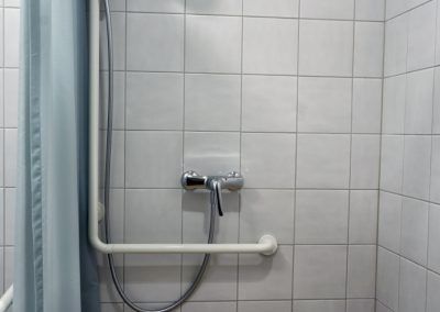 Dusche mit weiß gefliesten Wänden, Chromhahn, Haltegriff und blauem Duschvorhang.