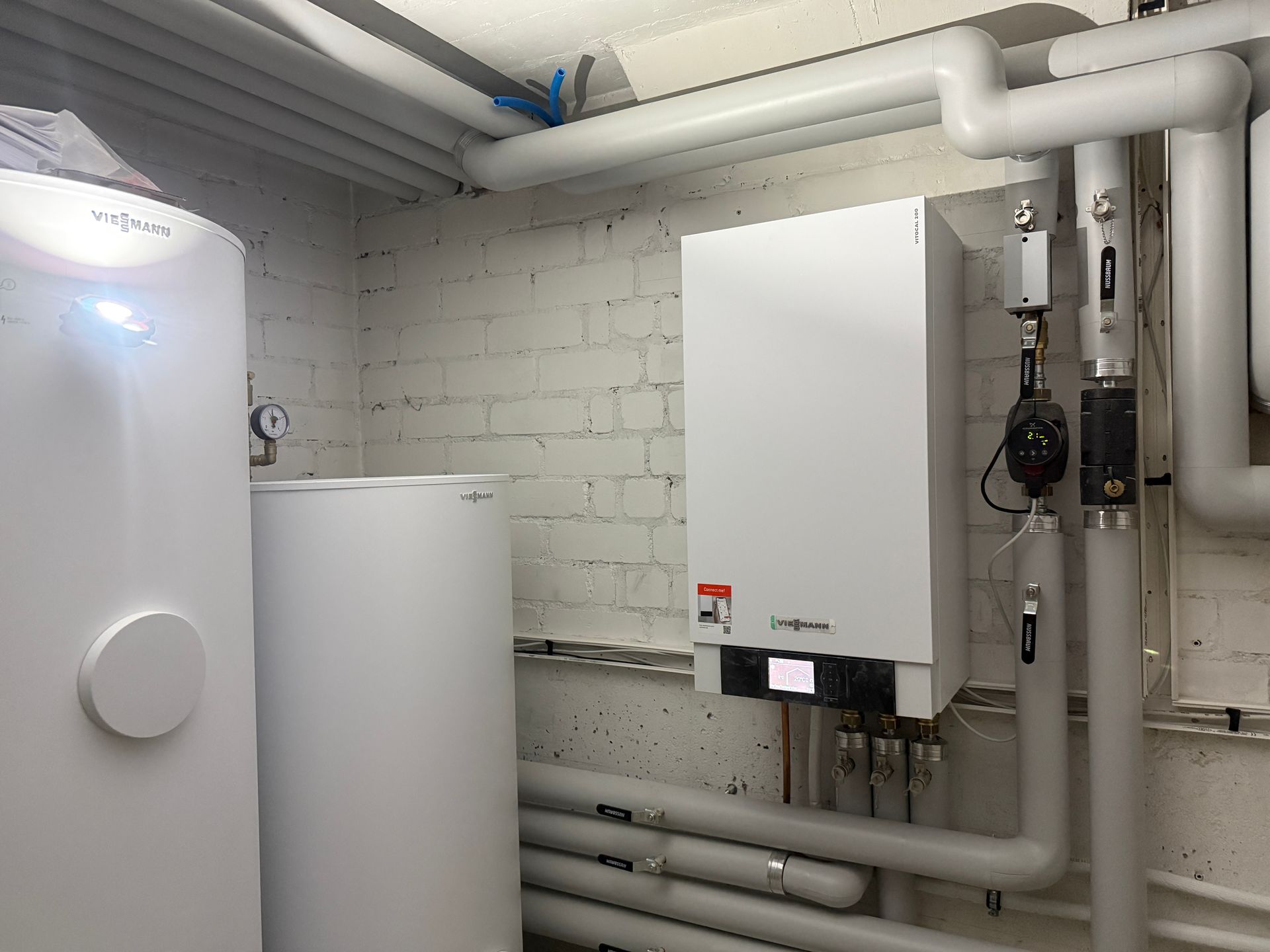 Eine moderne, weiße Heizungsanlage in einem kleinen Raum mit Kupferrohren, einem grünen Druckbehälter und einem grauen Boden.