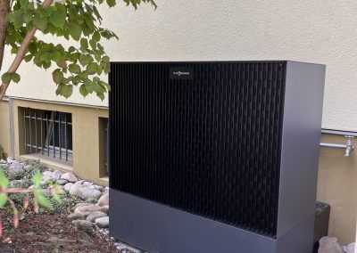 Große schwarze Wärmepumpeneinheit neben einem cremefarbenen Gebäude und einigen Pflanzen im Freien.