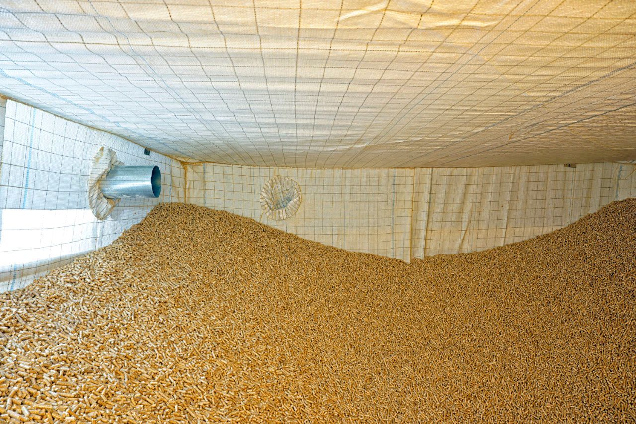 Ein Stapel Holzpellets füllt einen Lagerraum, in dem eine Metallentlüftung sichtbar ist.