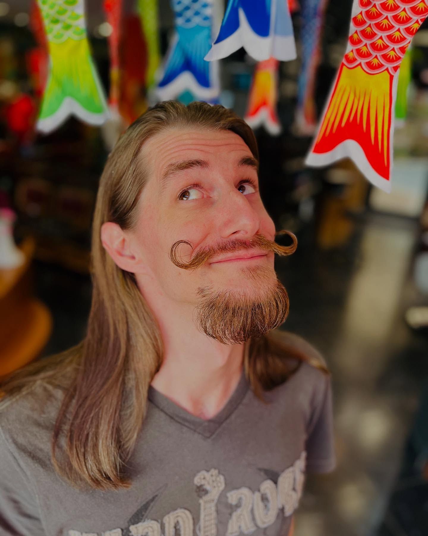 Une personne aux cheveux longs et à la moustache en guidon sourit vers le ciel, encadrée par des décorations de poissons en papier colorées suspendues.