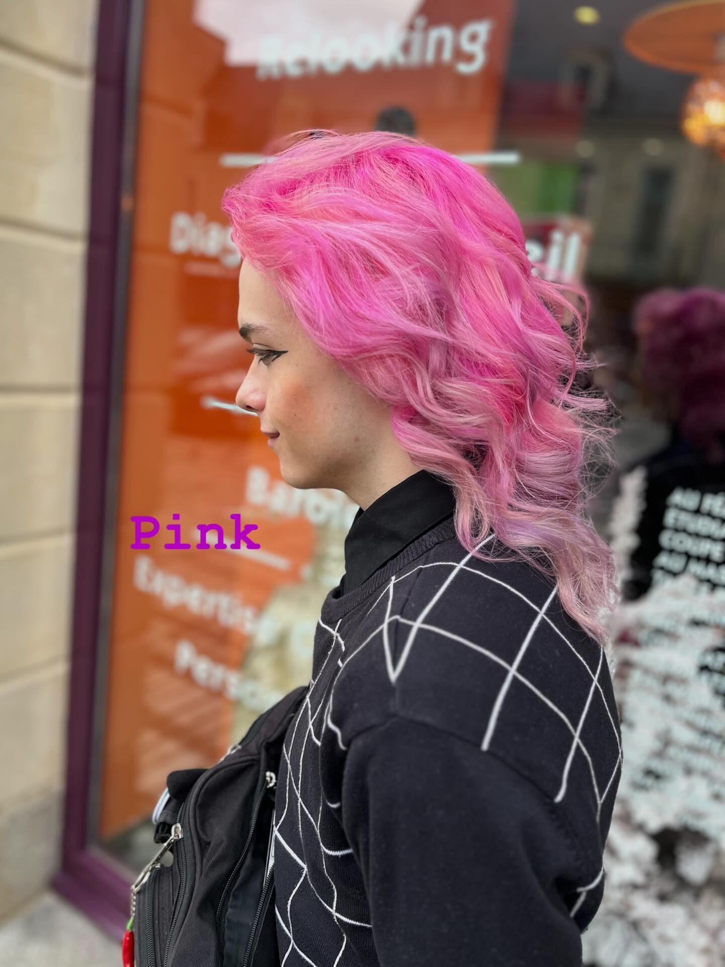 Une personne aux cheveux ondulés rose vif, mi-longs, se tient de profil devant la vitrine d'un salon de coiffure.