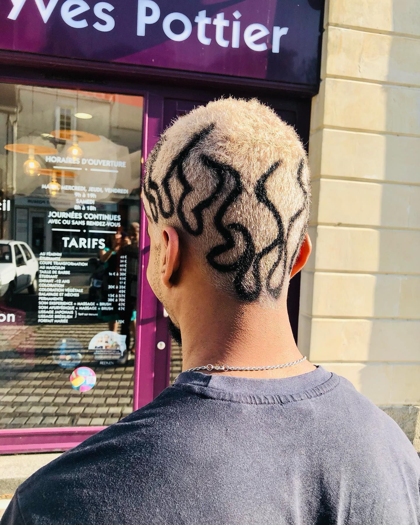 Une personne aux cheveux courts, blond platine, ornée d'une coiffure foncée en forme de flamme, aperçue devant un salon de coiffure.