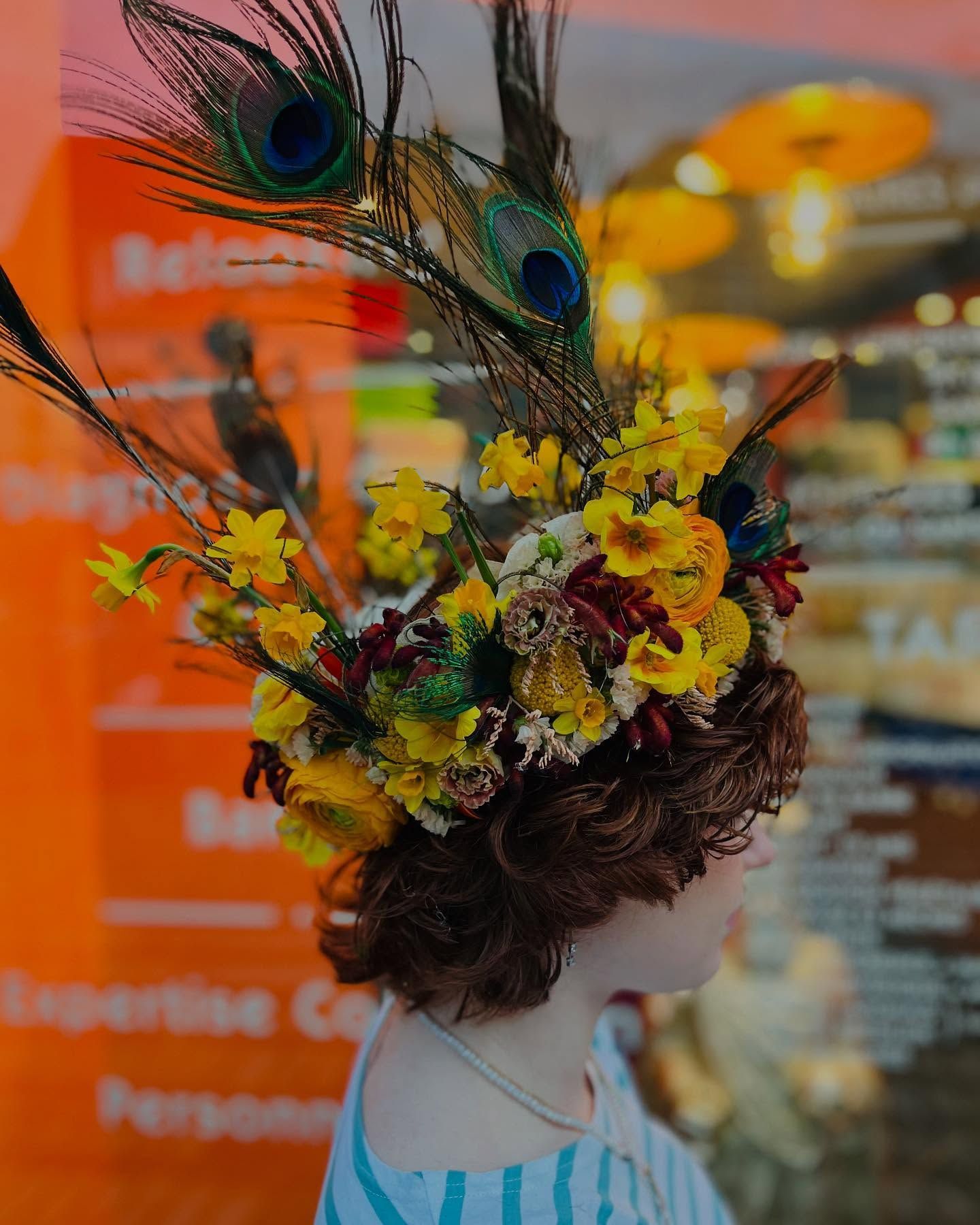 Une personne aux cheveux rose vif porte une couronne de fleurs et une chemise à rayures, souriant devant une devanture de magasin orange.