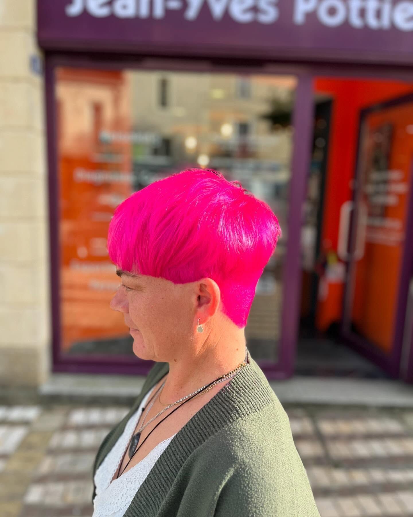 Une personne aux cheveux rose vif et aux lunettes à monture rouge sourit derrière un grand bouquet de jonquilles jaunes.