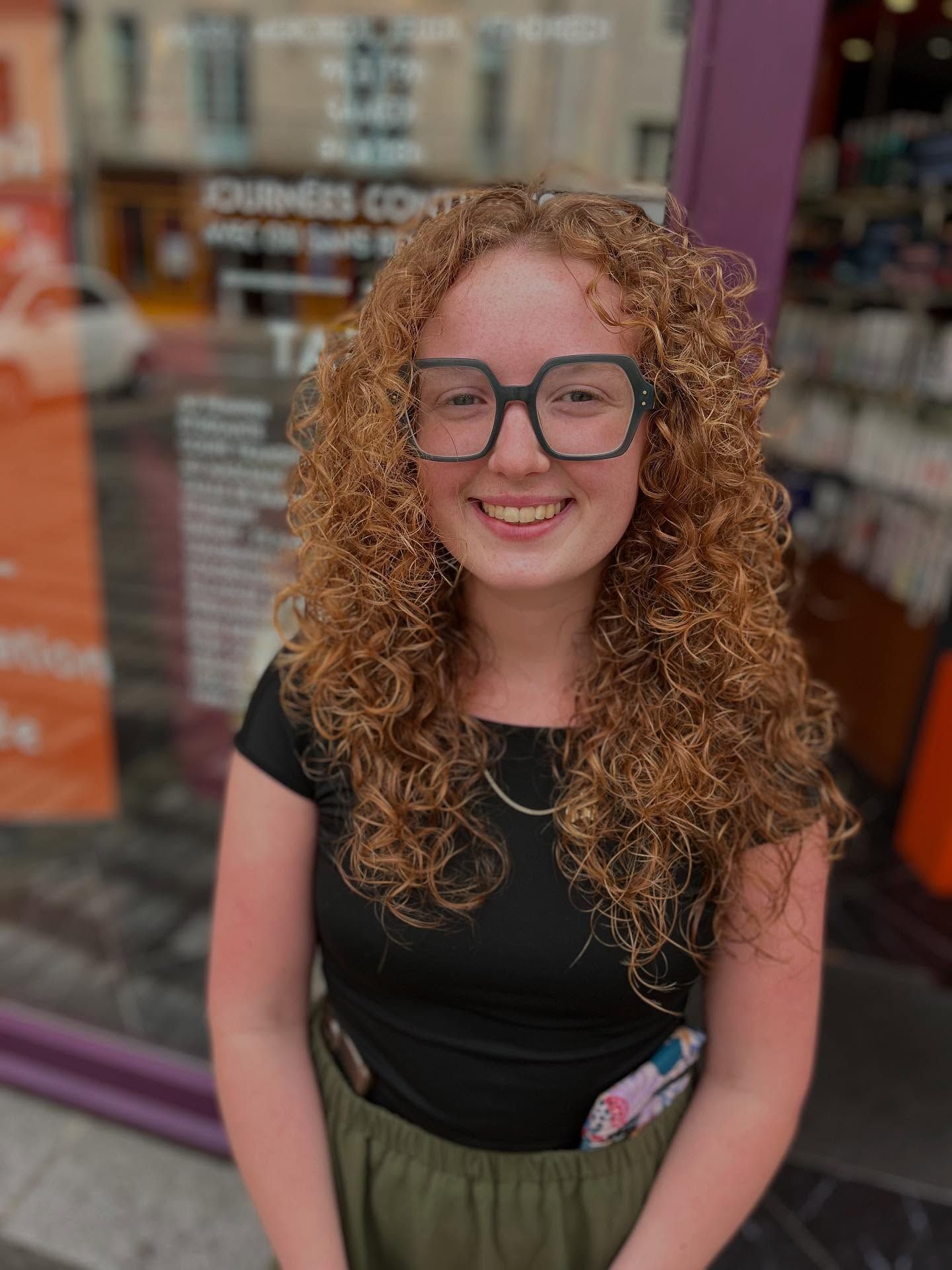 Personne souriante aux longs cheveux bouclés auburn et portant des lunettes vert foncé à monture carrée, vêtue d'un haut noir à l'extérieur.