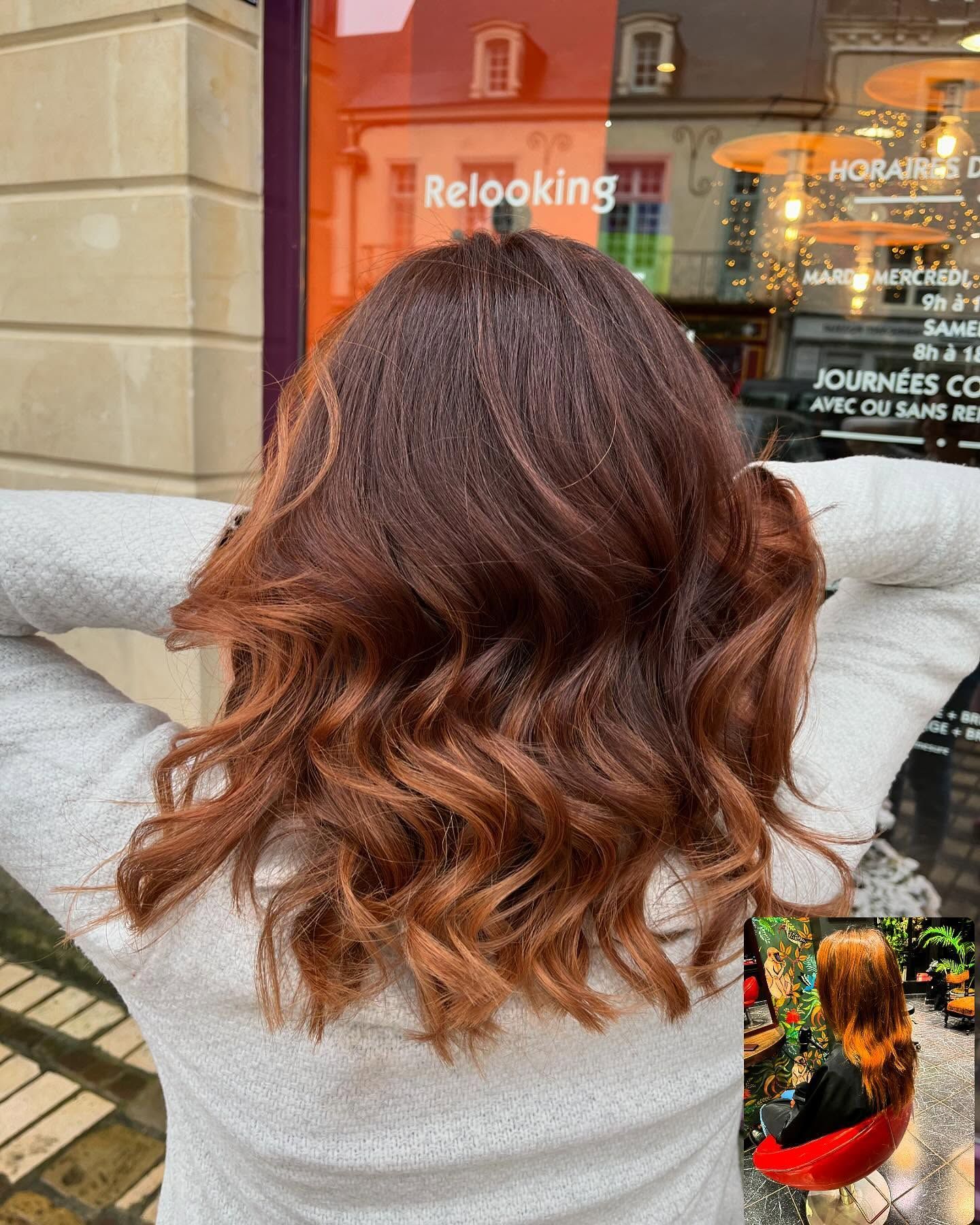 Une personne aux cheveux châtain clair ondulés, mi-longs, avec des reflets cuivrés, vue de dos dans un salon de coiffure.