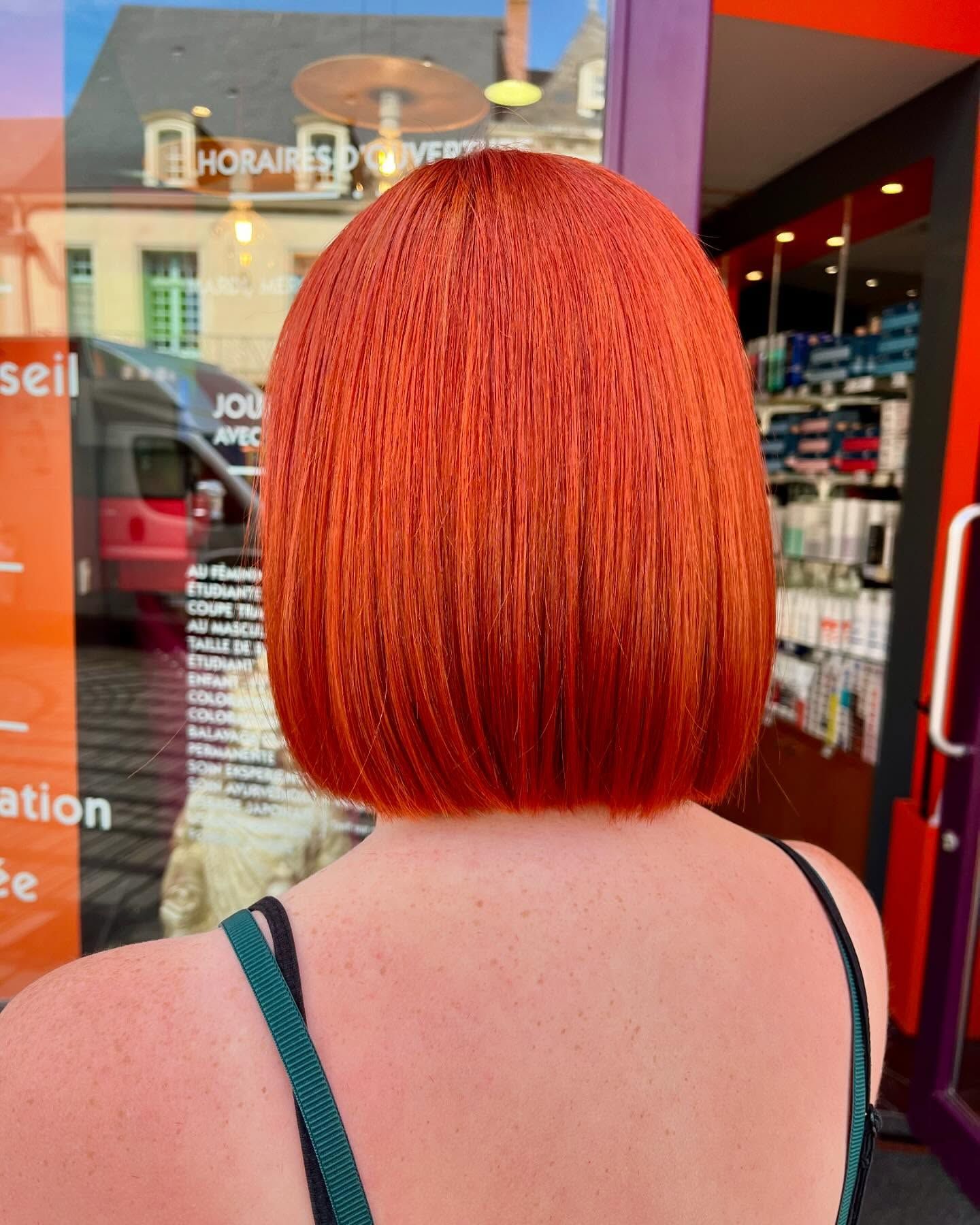 Une personne aux cheveux roux courts et ondulés, portant des lunettes et une chemise blanche à imprimé botanique orange et vert.