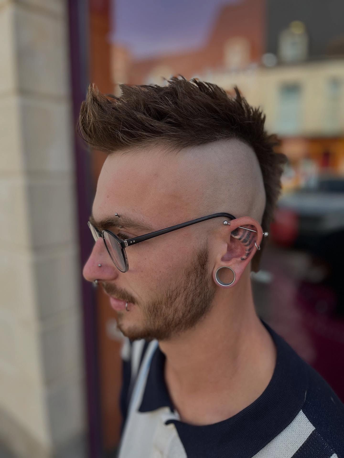 Profil latéral d'une personne avec une crête iroquoise, les côtés rasés, des lunettes et plusieurs piercings aux oreilles, en extérieur.