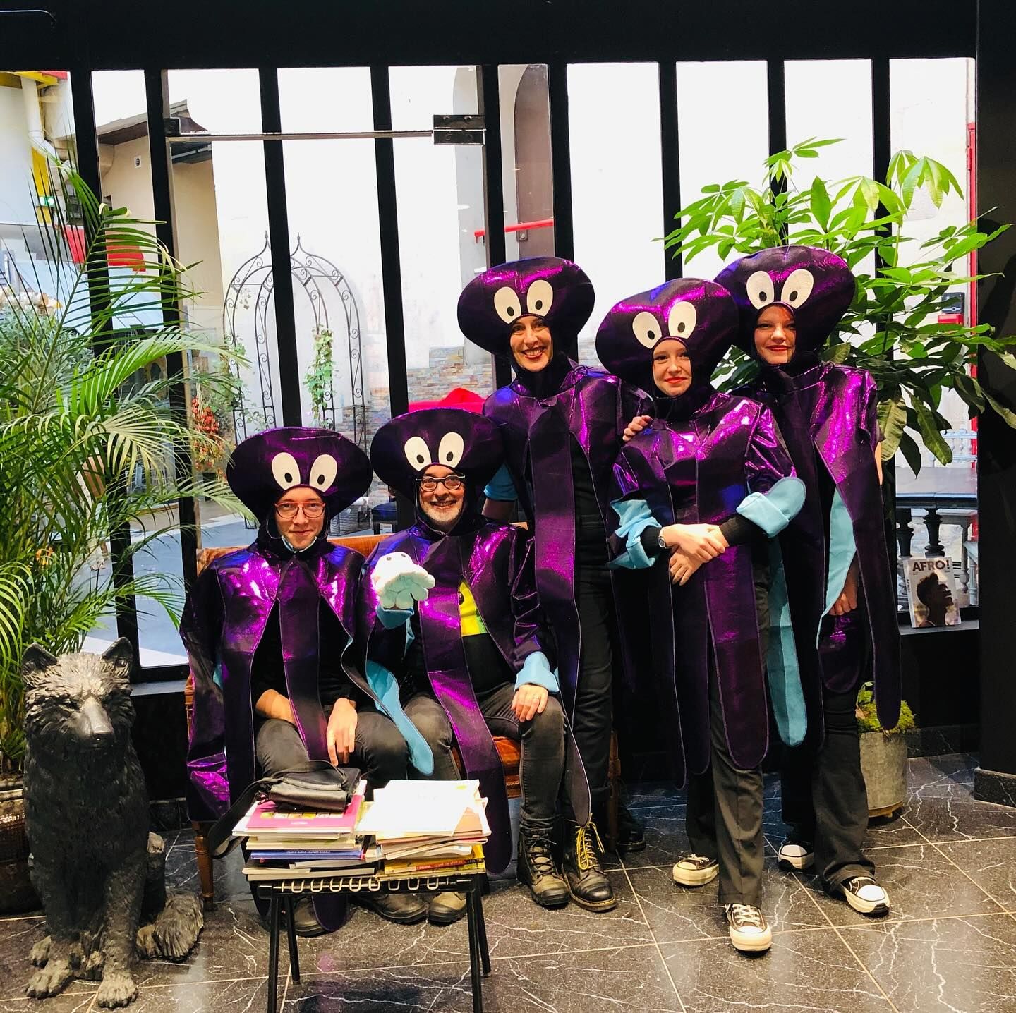 Cinq personnes vêtues de costumes et de coiffes assortis, de couleur violette, sur le thème des pieuvres, posent ensemble à l'intérieur, près d'une table et d'une statue.