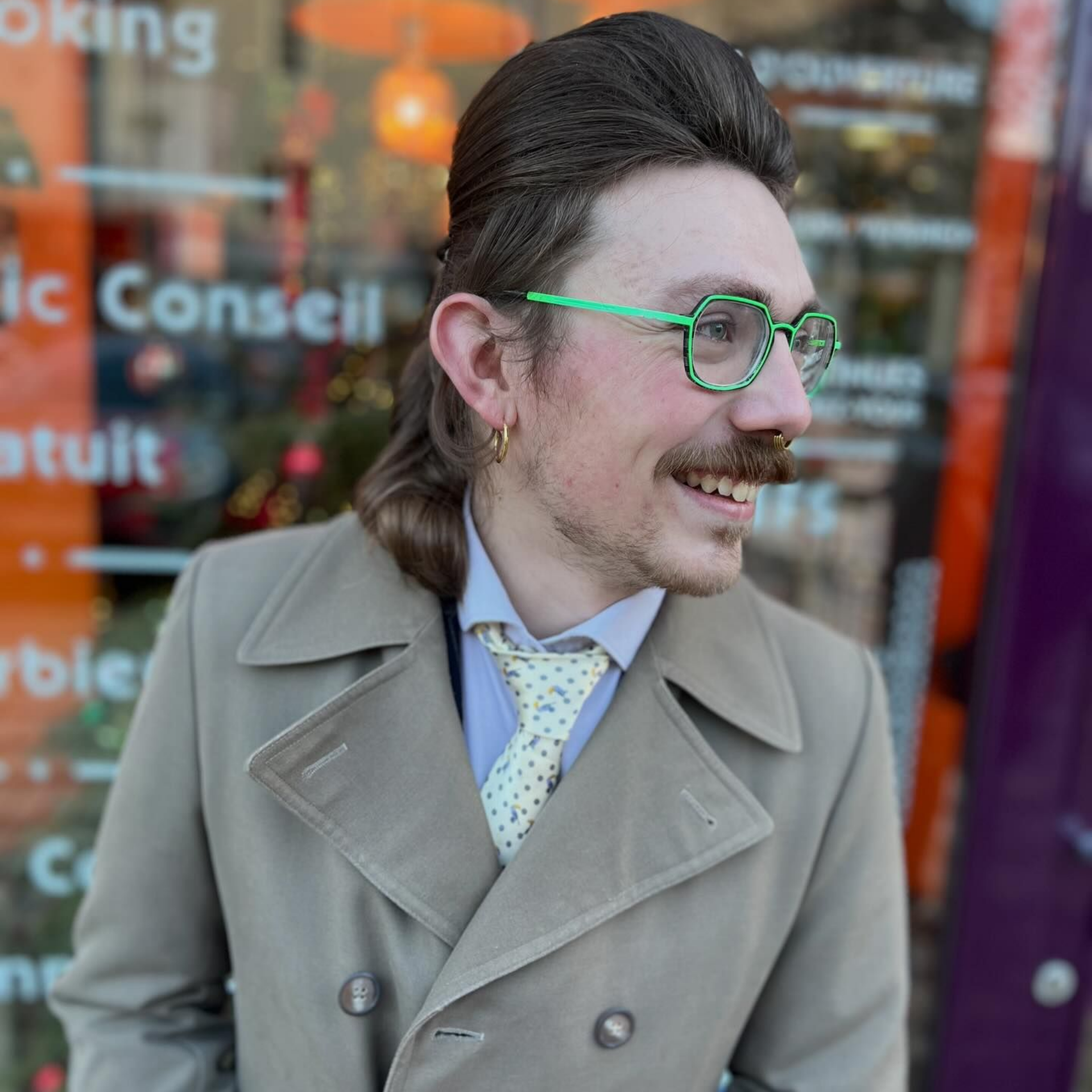 Une personne avec une coupe mulet brune, portant un manteau beige, une cravate à motifs clairs et des lunettes vert vif, souriant à l'extérieur.