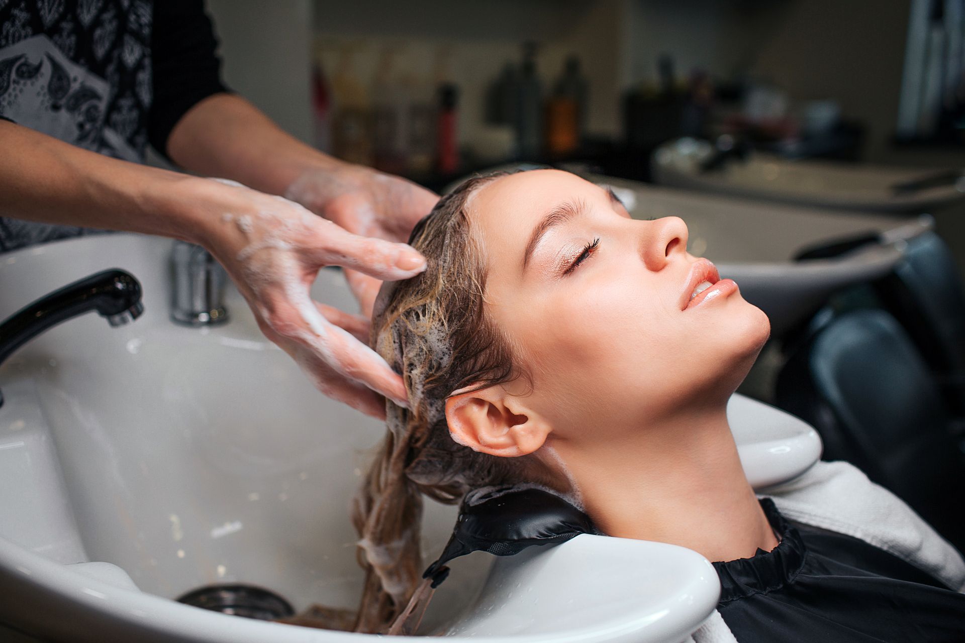Une coiffeuse lave les cheveux d'une cliente au lavabo du salon ; la cliente a les yeux fermés et une expression détendue.