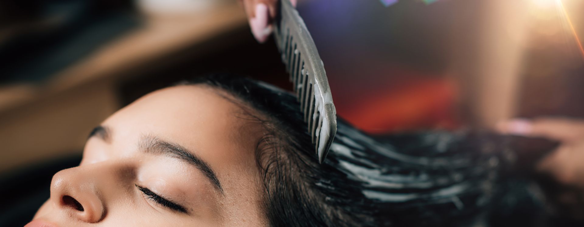 Une personne, les yeux fermés, reçoit un soin capillaire tandis qu'un coiffeur applique le produit sur ses cheveux à l'aide d'un peigne.