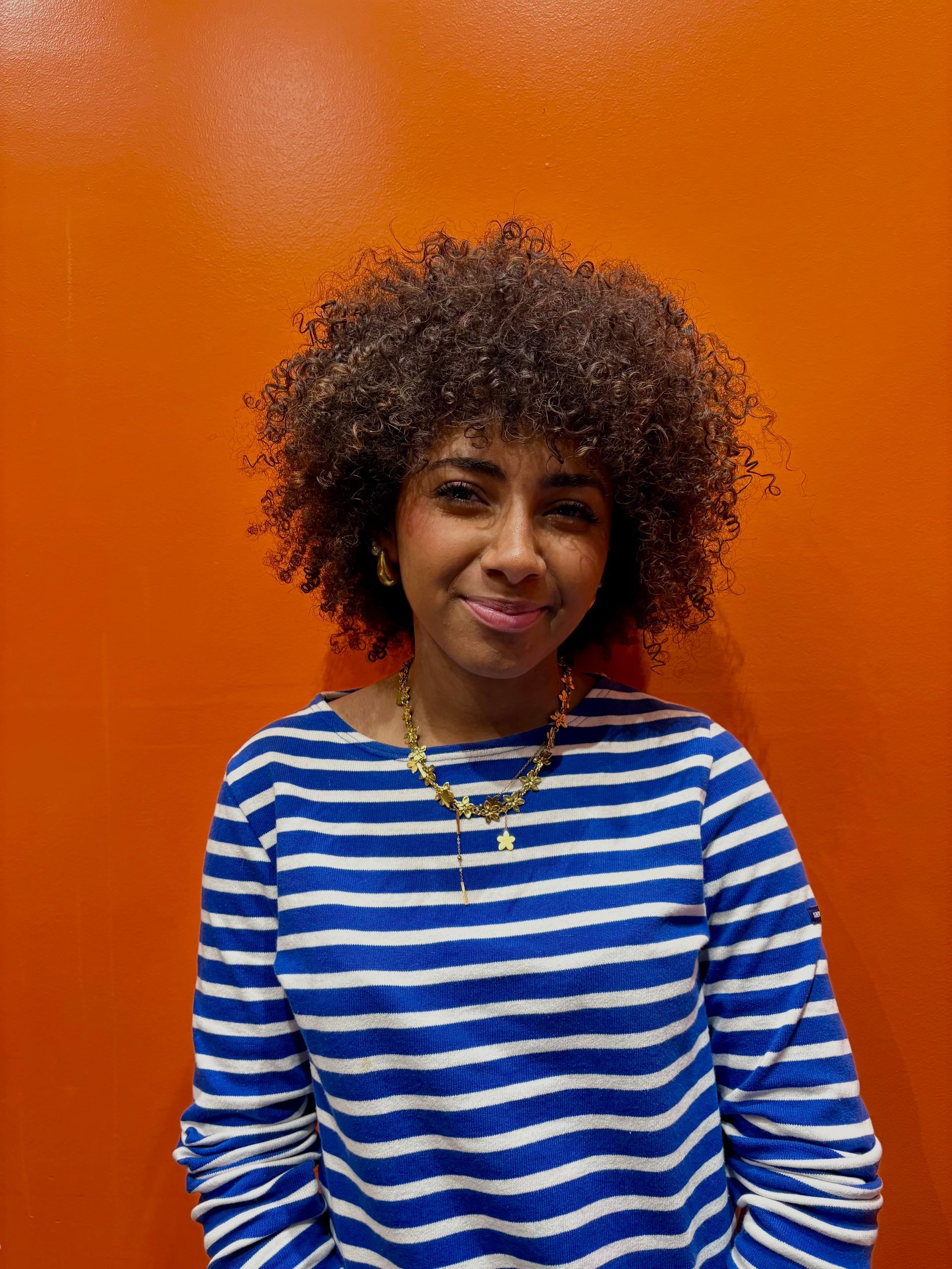 Une personne souriante aux cheveux bouclés porte une chemise à manches longues à rayures bleues et blanches sur un fond orange vif.