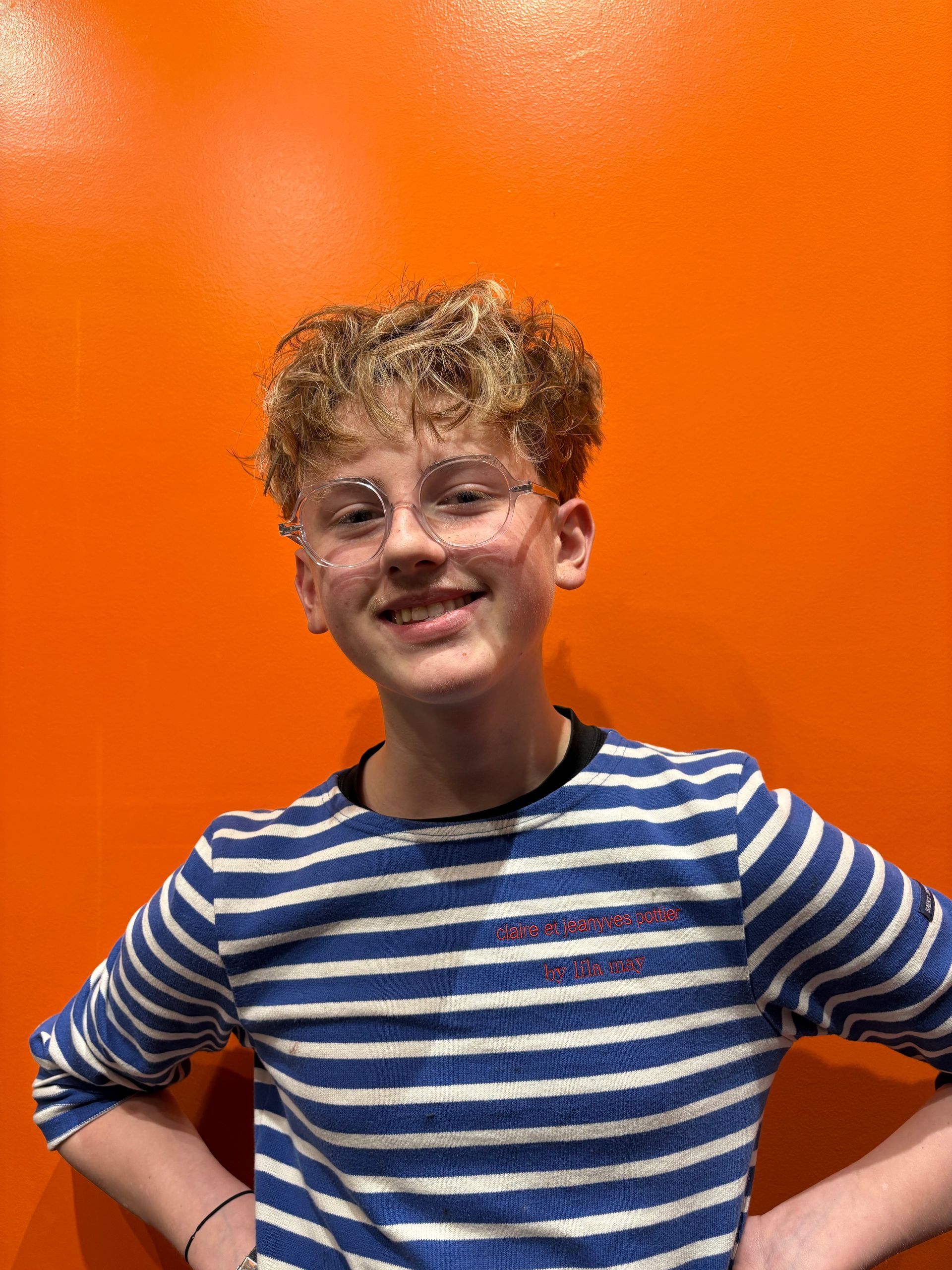 Une personne aux cheveux bouclés et portant des lunettes sourit, vêtue d'une chemise à rayures bleues et blanches, devant un mur orange vif.