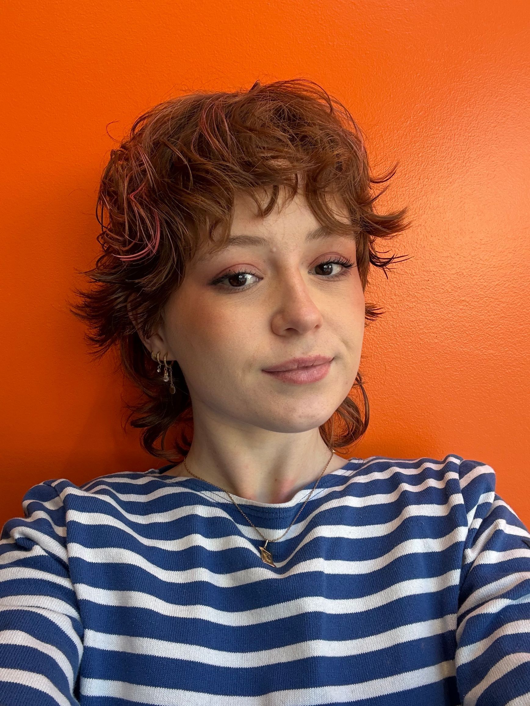 Une personne aux cheveux courts, bouclés et brun roux porte une chemise à rayures bleues et blanches sur un fond orange vif.