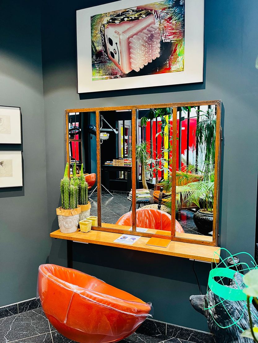 Un coin salon stylisé avec une étagère en bois, un grand miroir à plusieurs panneaux, une chaise orange vif et des plantes en pot.