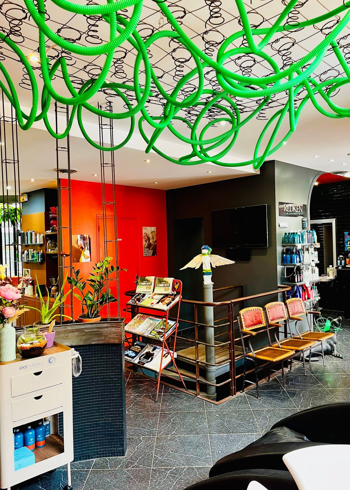 Un salon de coiffure avec une installation artistique en corde vert vif au plafond, un mur rouge et une salle d'attente.