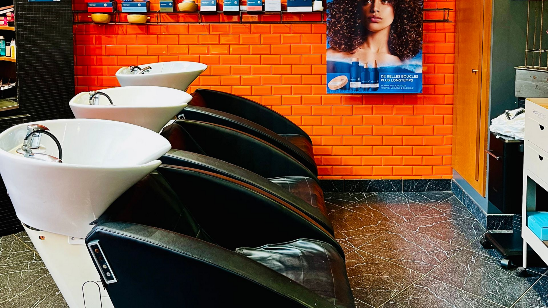 Une rangée de trois bacs à shampoing de salon contre un mur de briques orange vif, avec une affiche au-dessus.