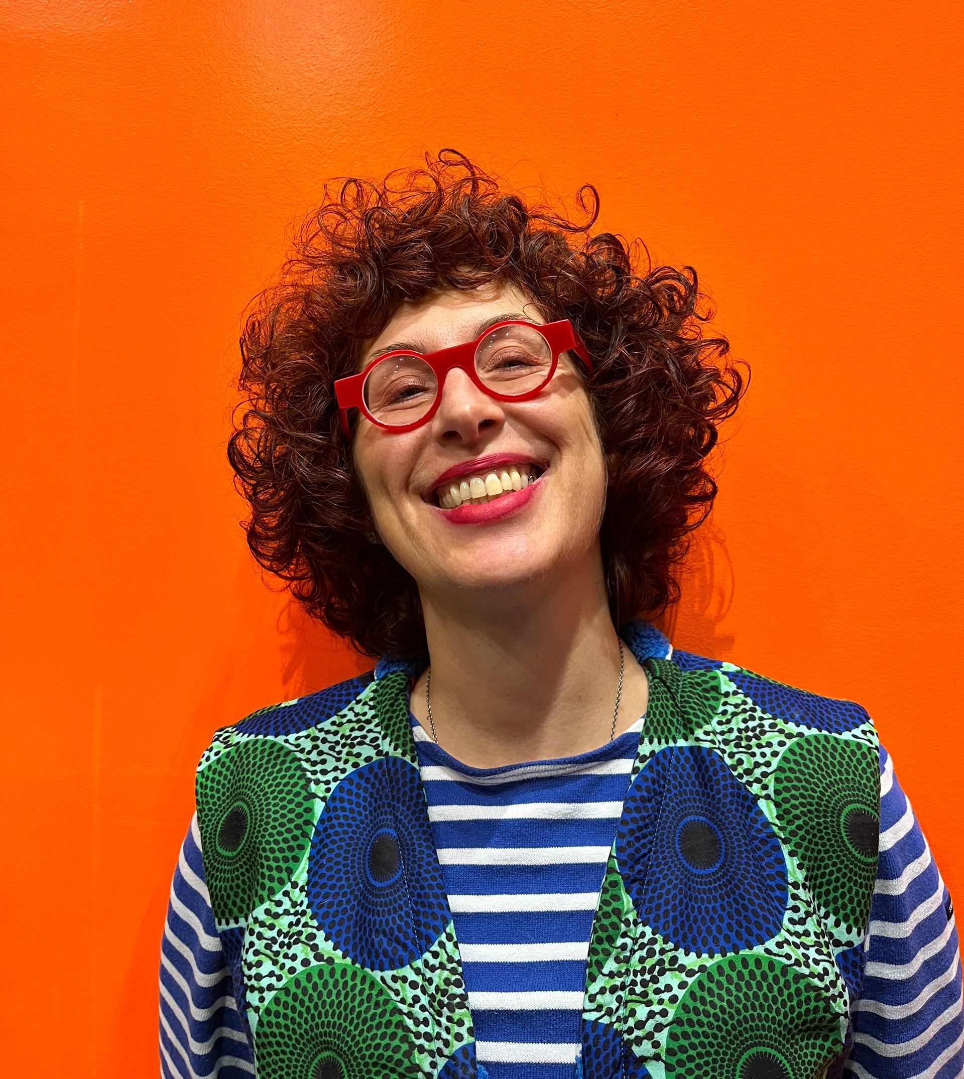 Une personne aux cheveux bouclés et aux lunettes rouges sourit sur un fond orange vif, vêtue d'une chemise à rayures et d'un gilet à motifs.