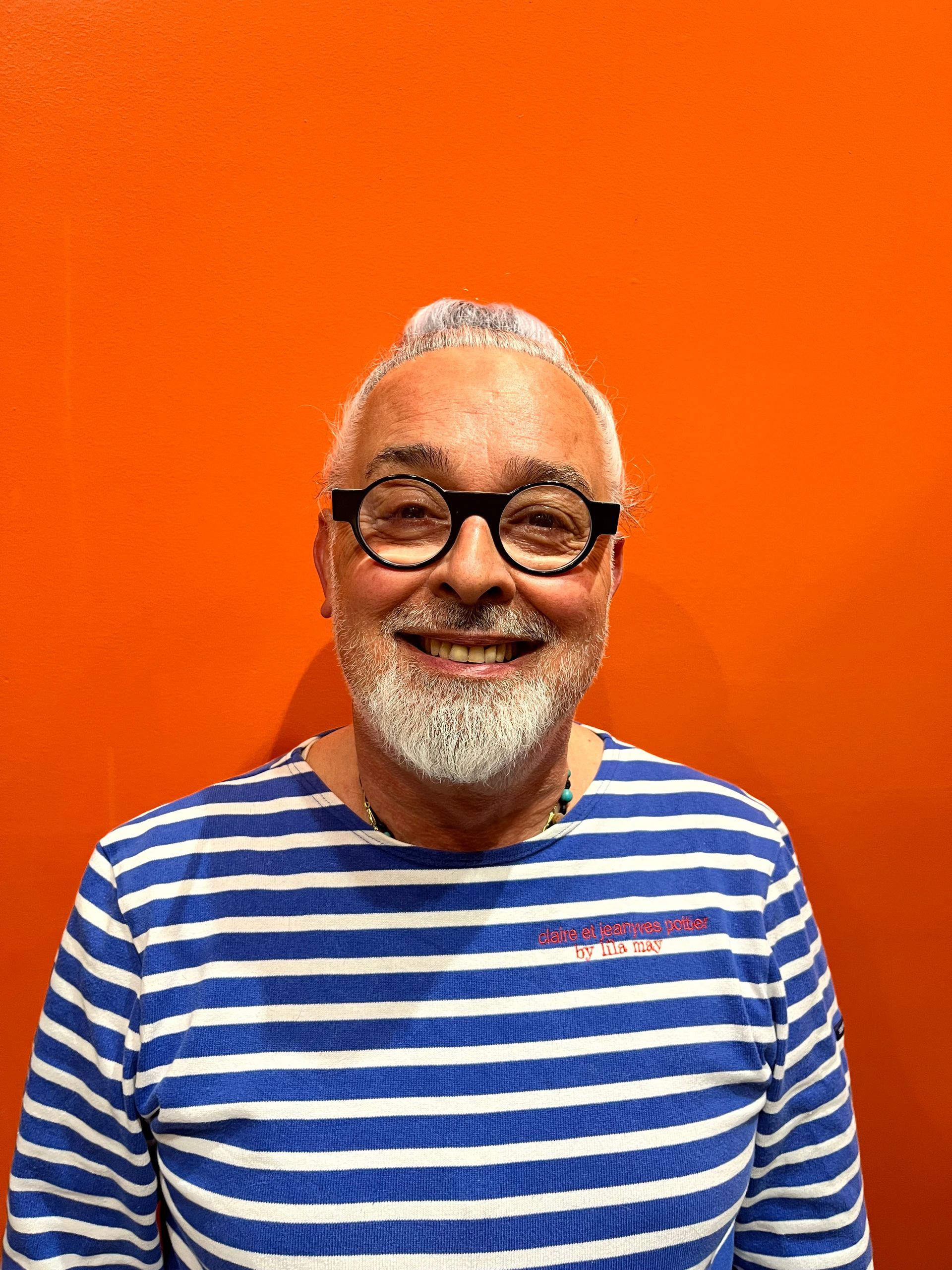 Personne souriante aux cheveux gris courts, à la barbe fournie et portant des lunettes noires, vêtue d'une chemise à rayures bleues et blanches sur fond orange.